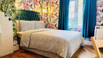 Een slaapkamer met bloemenbehang, een groen gestoffeerd hoofdeinde, een bed met beddengoed met een patroon, en een raam met blauwe gordijnen.