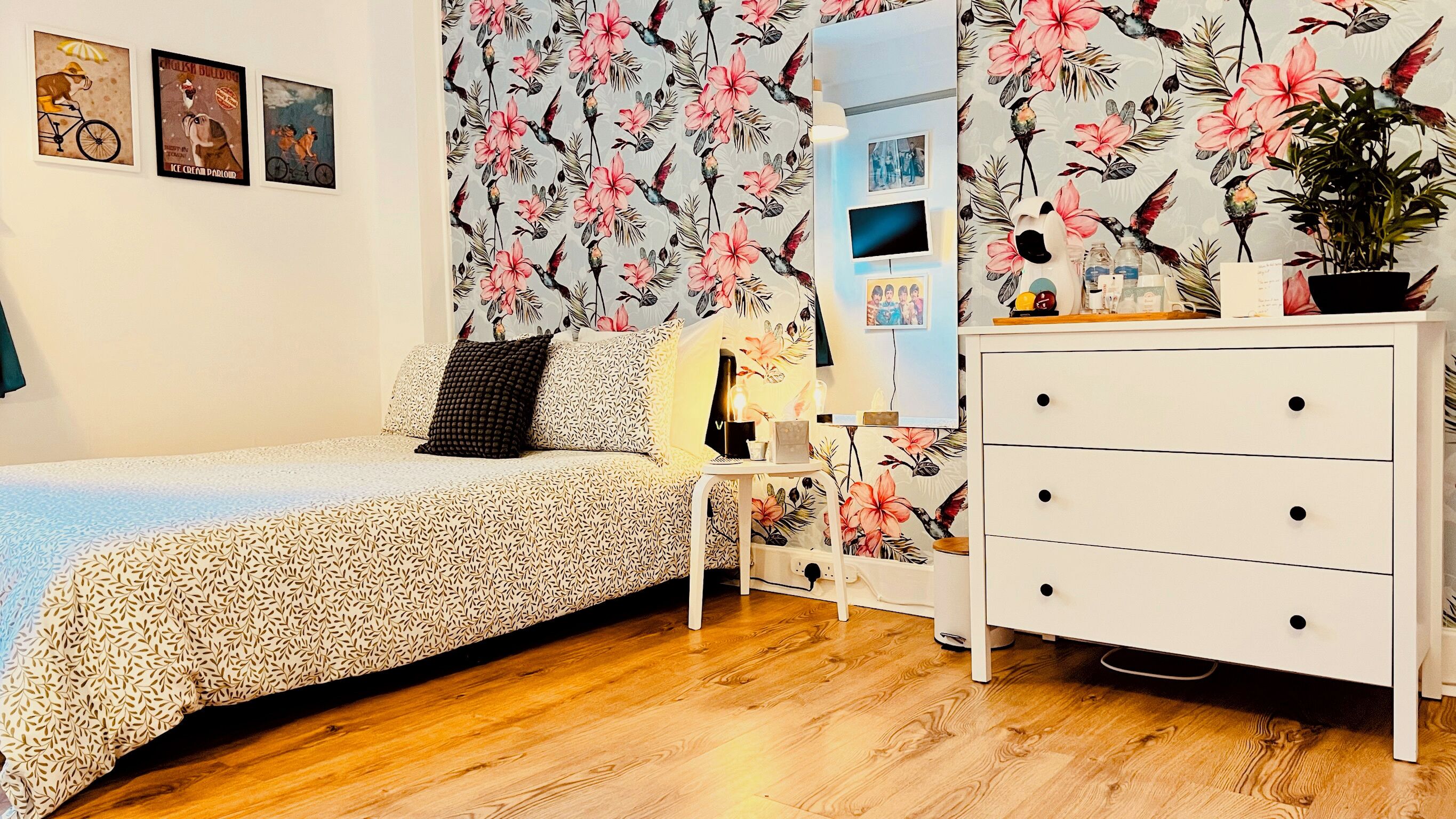 A bedroom with a floral wallpaper, a bed with a patterned duvet, a white dresser, and a small table with a lamp.