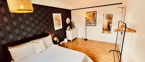 A modern bedroom with a patterned bedspread, a hanging light fixture, a dresser, and artwork on the walls.