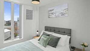 Ein Schlafzimmer mit großem Fenster, einem Bett mit grau-weißer Bettwäsche, einem grauen Kopfteil, einem Nachttisch mit Lampe und einem Gemälde an der Wand.