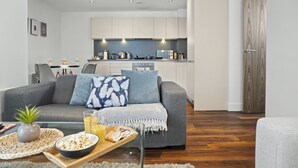 Ein modernes Wohnzimmer mit einem grauen Sofa, einem Couchtisch mit Snacks und einer Küche im Hintergrund.