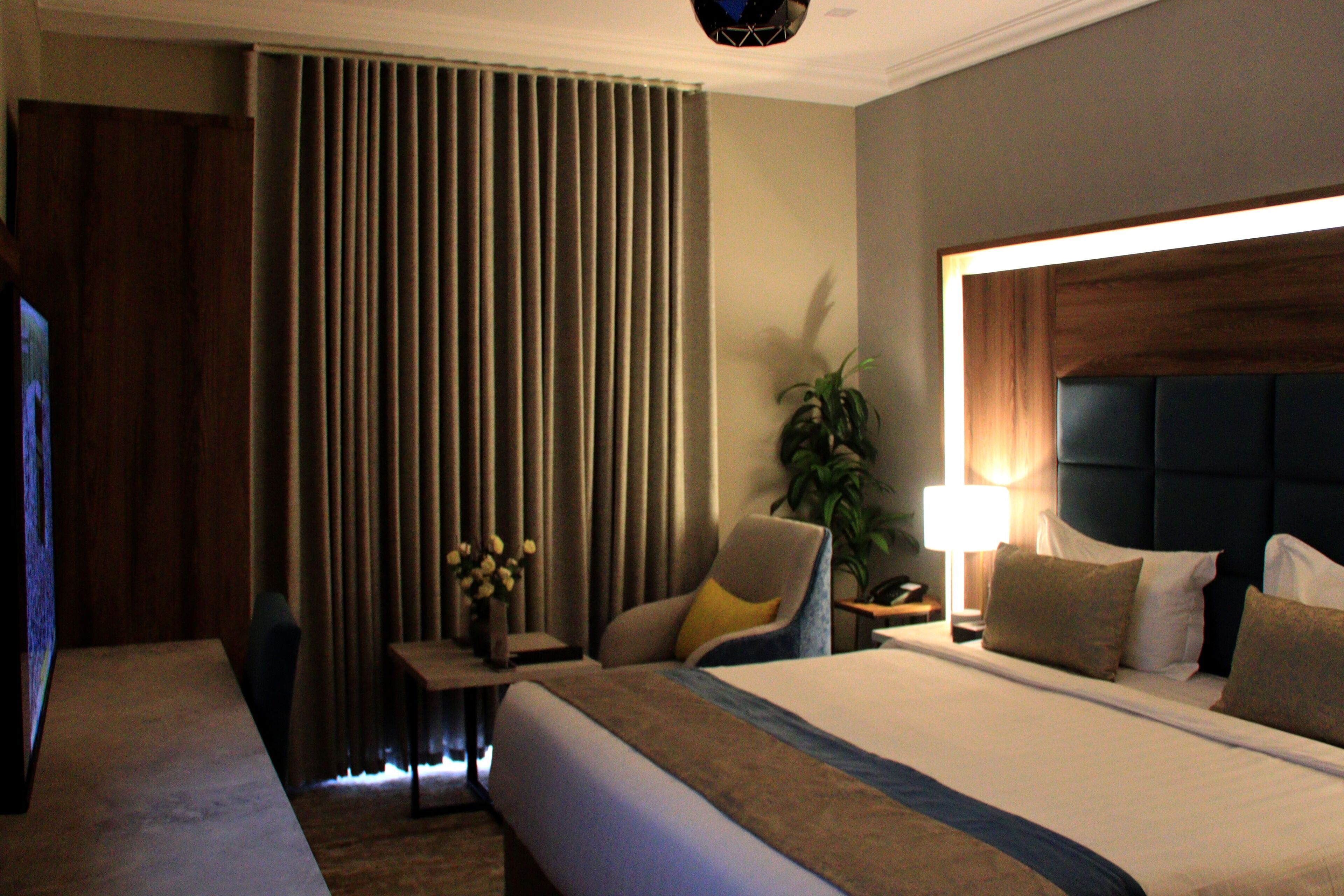 A hotel room with a large bed, a desk with a chair, a TV, a lamp, and a plant.