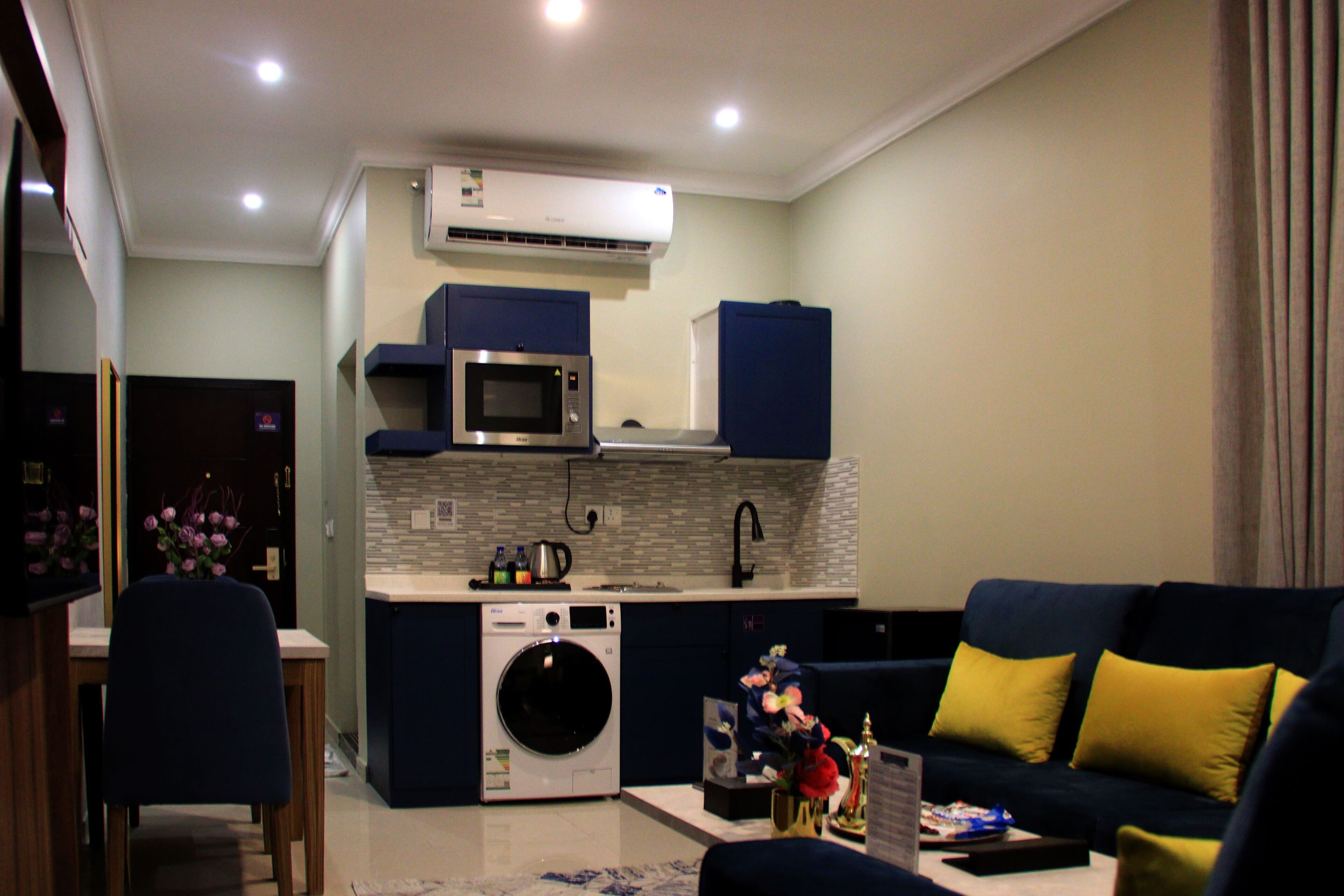 A compact living space with a kitchenette, a washing machine, a sofa with yellow cushions, and a dining area with a chair and table.
