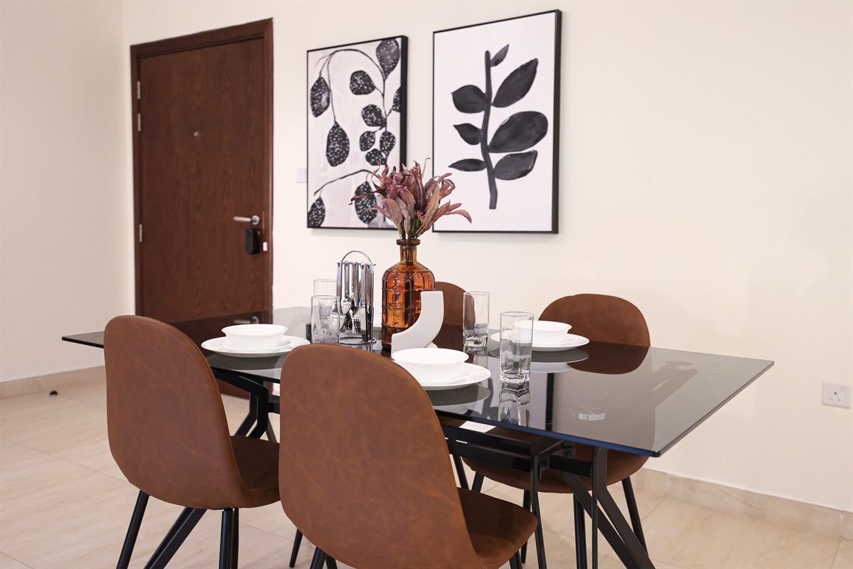 Un coin repas avec une table en verre, quatre chaises marron, un vase de fleurs et deux reproductions botaniques encadrées accrochées au mur.