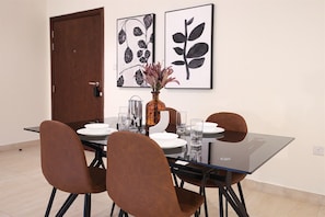 A dining area with a glass table, four brown chairs, a vase with flowers, and two framed botanical prints on the wall. - Stylish 1BR in Azizi Iris, Al Furjan (Dubai)