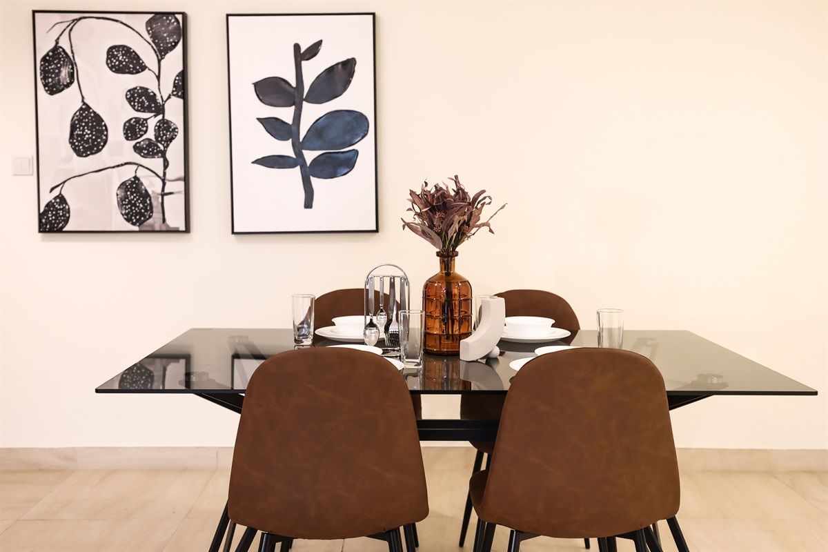 Ein Essbereich mit einem Glastisch, vier braunen Stühlen, einer Vase mit getrockneten Blumen und zwei gerahmten Kunstwerken an der Wand.