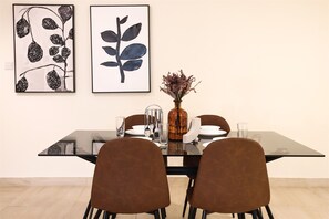 A dining area with a glass table, four brown chairs, a vase with dried flowers, and two framed artworks on the wall. - Stylish 1BR in Azizi Iris, Al Furjan (Dubai)
