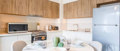 A modern kitchen with wooden cabinets, a stainless steel refrigerator, a microwave, a stove, and a dining table set for four.