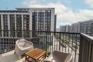 A balcony with a sofa, a wooden table, and a view of modern buildings. - Upscale & Stylish 2BR Dubai Hills (Dubai)