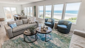 A spacious living room with a large window overlooking the beach, a grey sofa, a blue armchair, and a coffee table with decorative items. - Beach Me! Oceanfront steps2sand (Lincoln City)