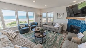 A spacious living room with a large window overlooking the beach, a fireplace, and comfortable seating. - Beach Me! Oceanfront steps2sand (Lincoln City)