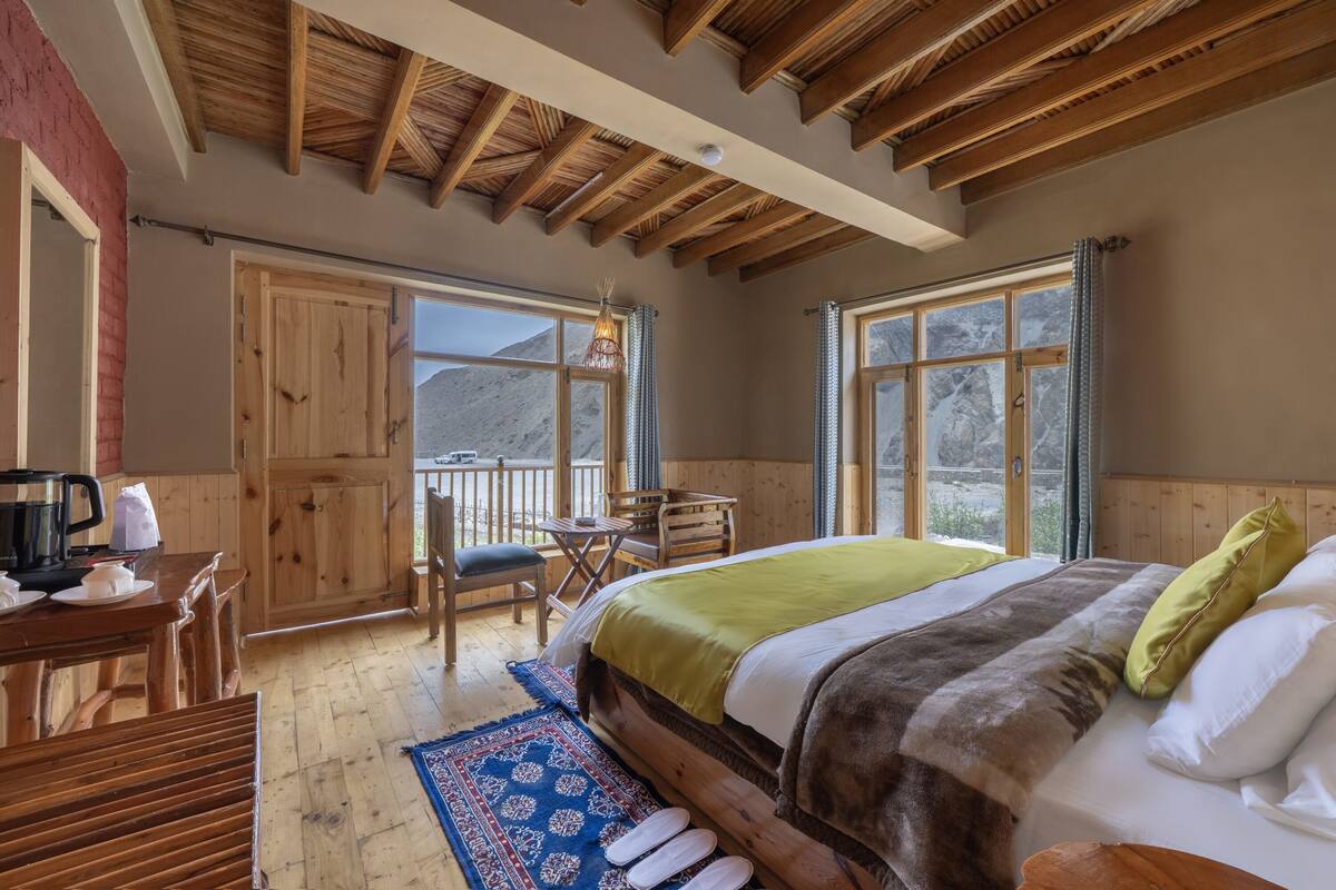 A wooden-framed room with a bed, a table with chairs, and a view of mountains through large windows.