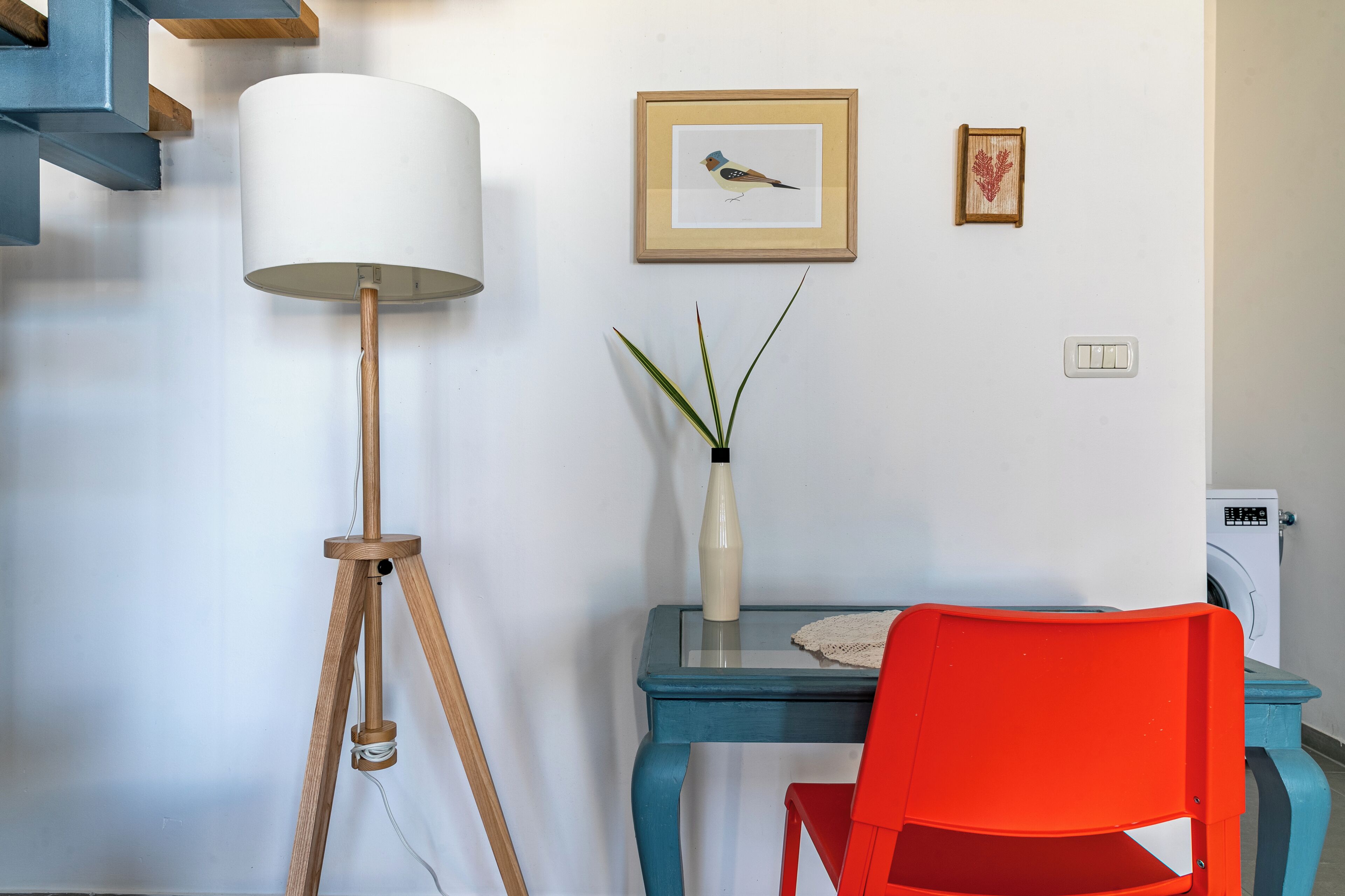 Une pièce comprenant un lampadaire sur trépied, une table avec un vase de plantes, un tableau encadré représentant un oiseau, une chaise rouge et un lave-linge.