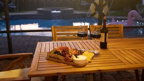 A wooden table with a cheese platter, two glasses of red wine, and a bottle of wine, set outdoors with a pool and a flamingo decoration in the background.