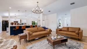 A spacious living room with two brown sofas, a coffee table, and a rug. A kitchen with a bar area is visible in the background.