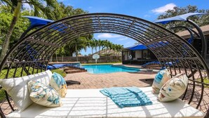 A sun lounger with cushions and a blue towel, a pool, and a covered seating area with umbrellas.
