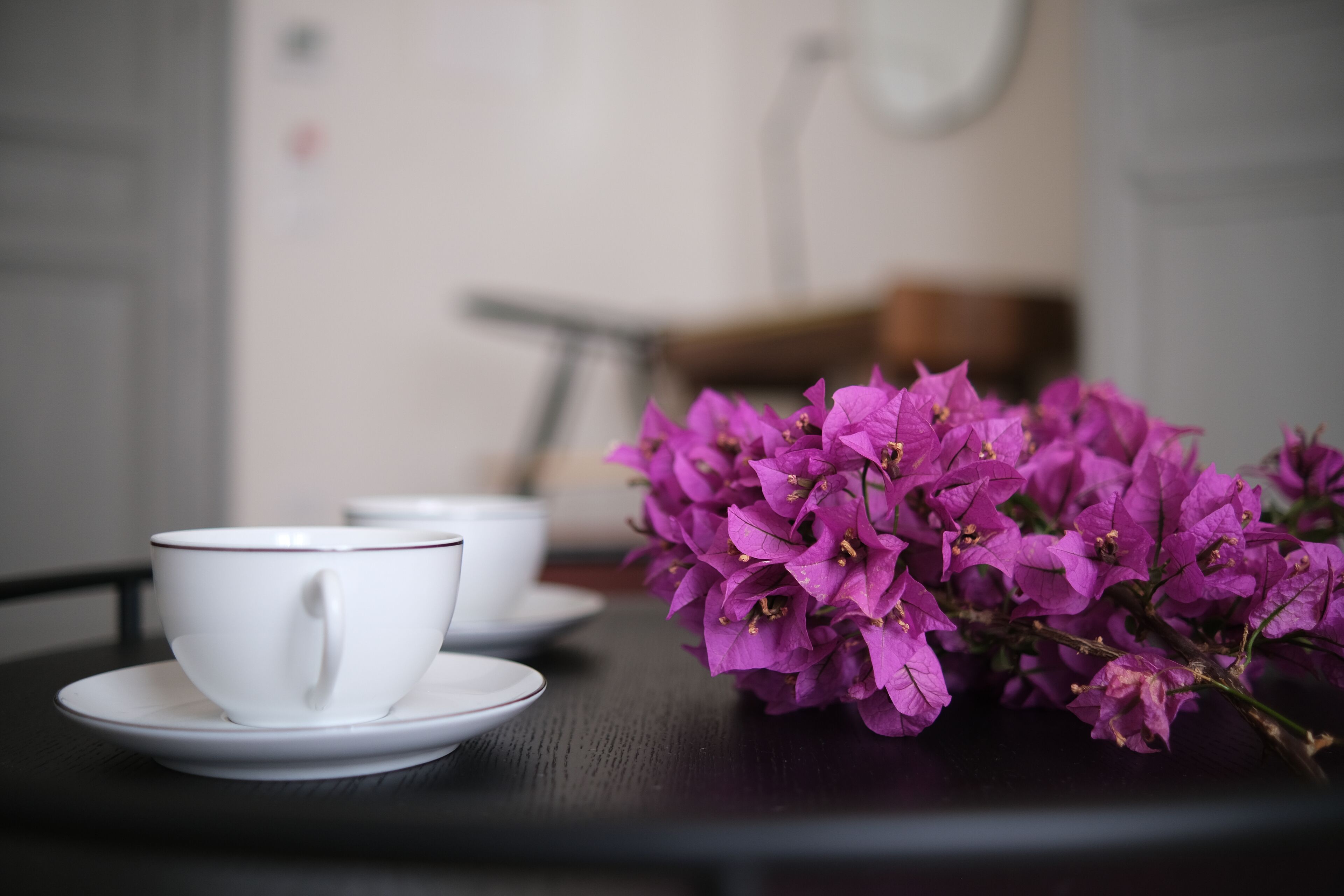 Ein Tisch mit zwei weißen Tassen und Untertellern sowie einem Strauß lila Blumen.
