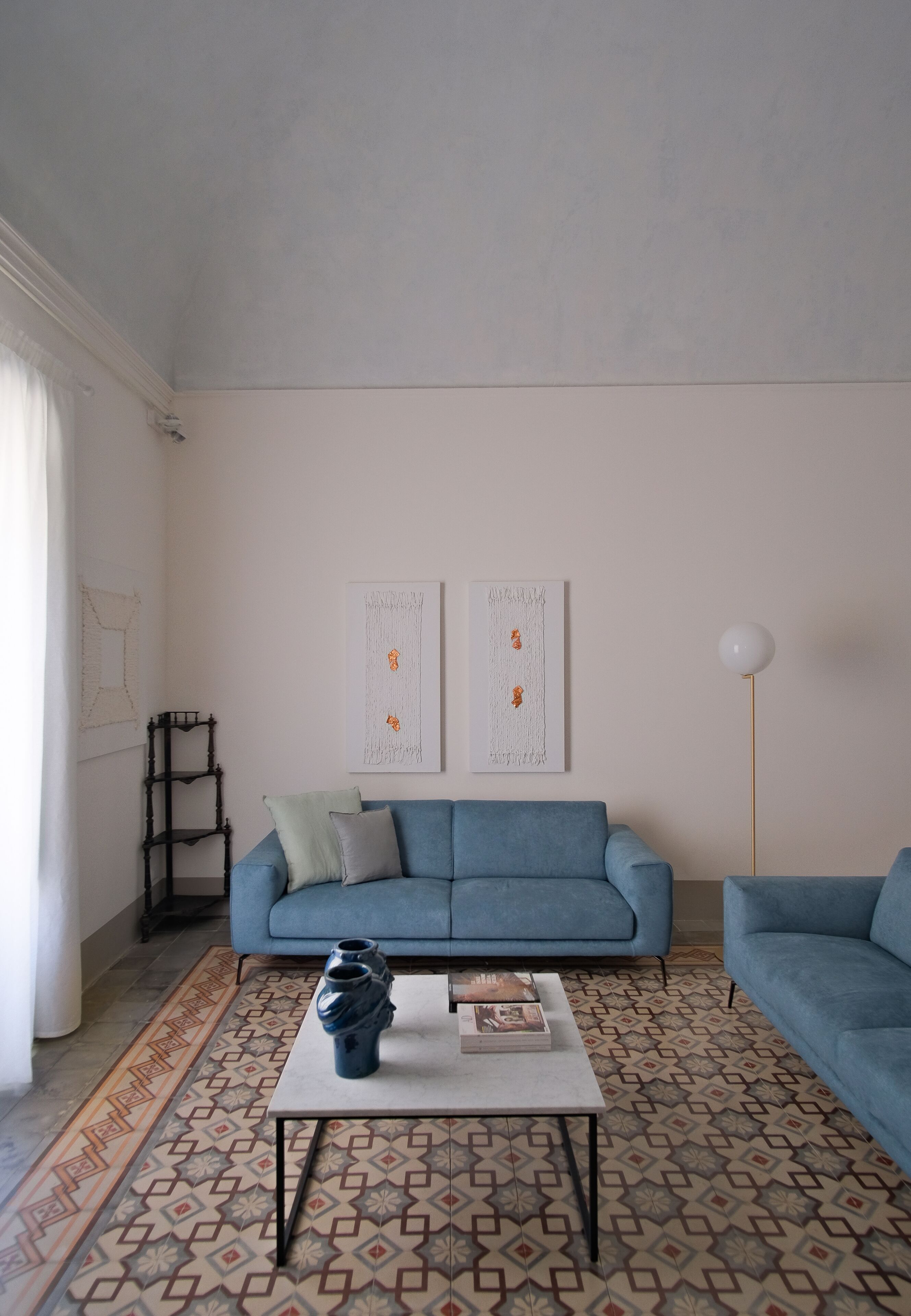 Een woonkamer met een blauwe bank, een salontafel en een kamerbreed tapijt. Er hangen twee kunstwerken aan de muur en staat een vloerlamp.