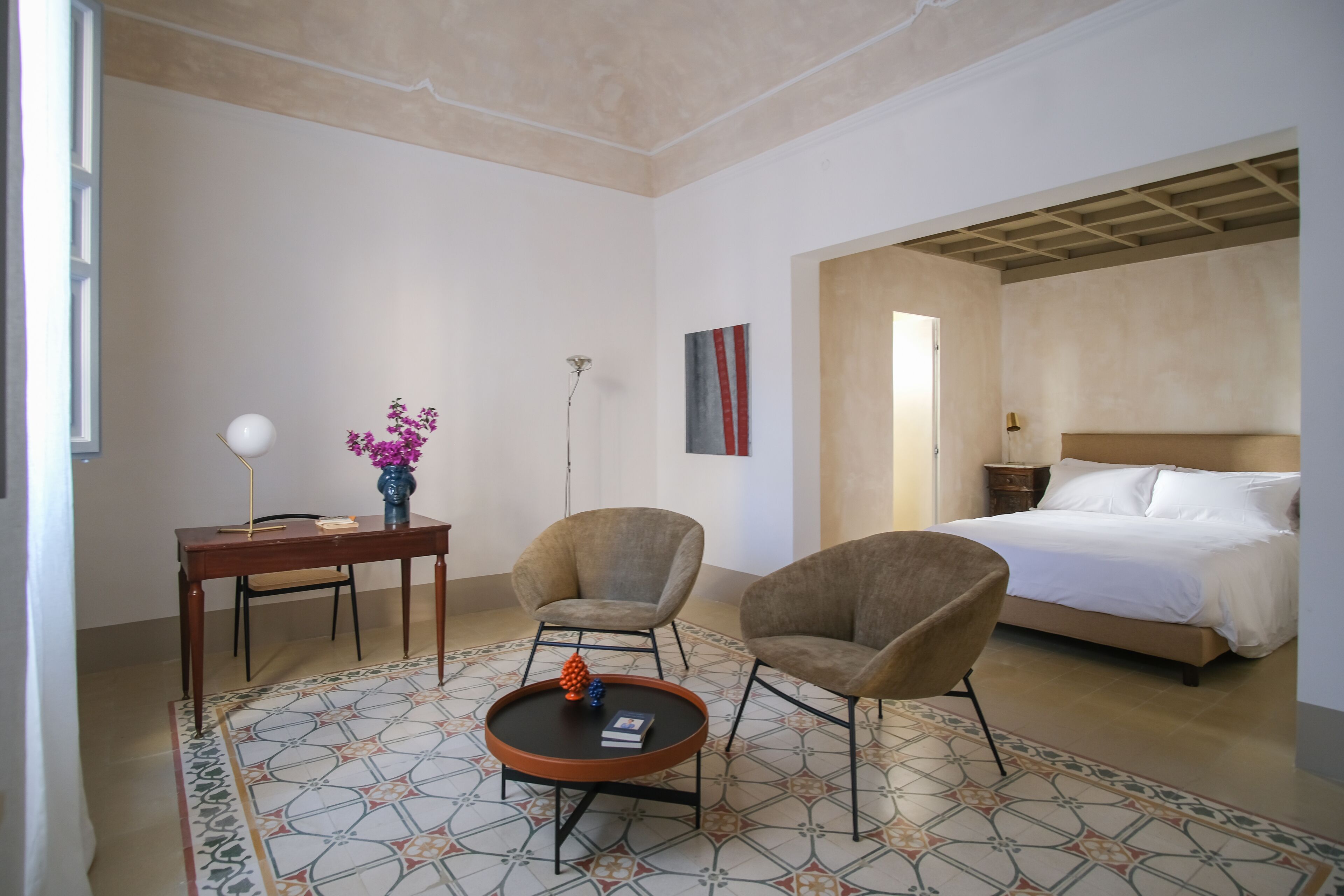 A hotel room with a bed, two armchairs, a small table, a lamp, and a vase with flowers.