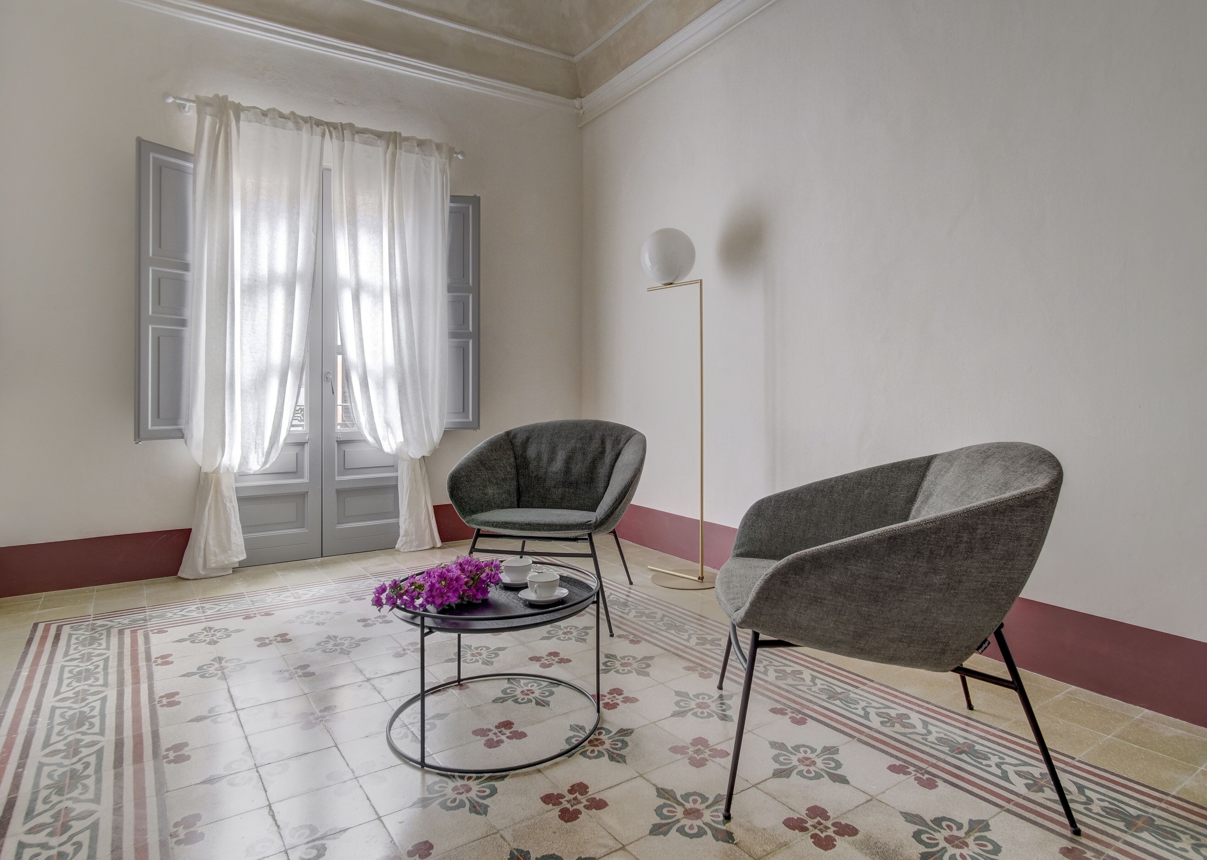Ein Zimmer mit zwei grauen Sesseln, einem kleinen runden Tisch mit einem lilafarbenen Blumenarrangement und einem Boden mit gemusterten Fliesen.