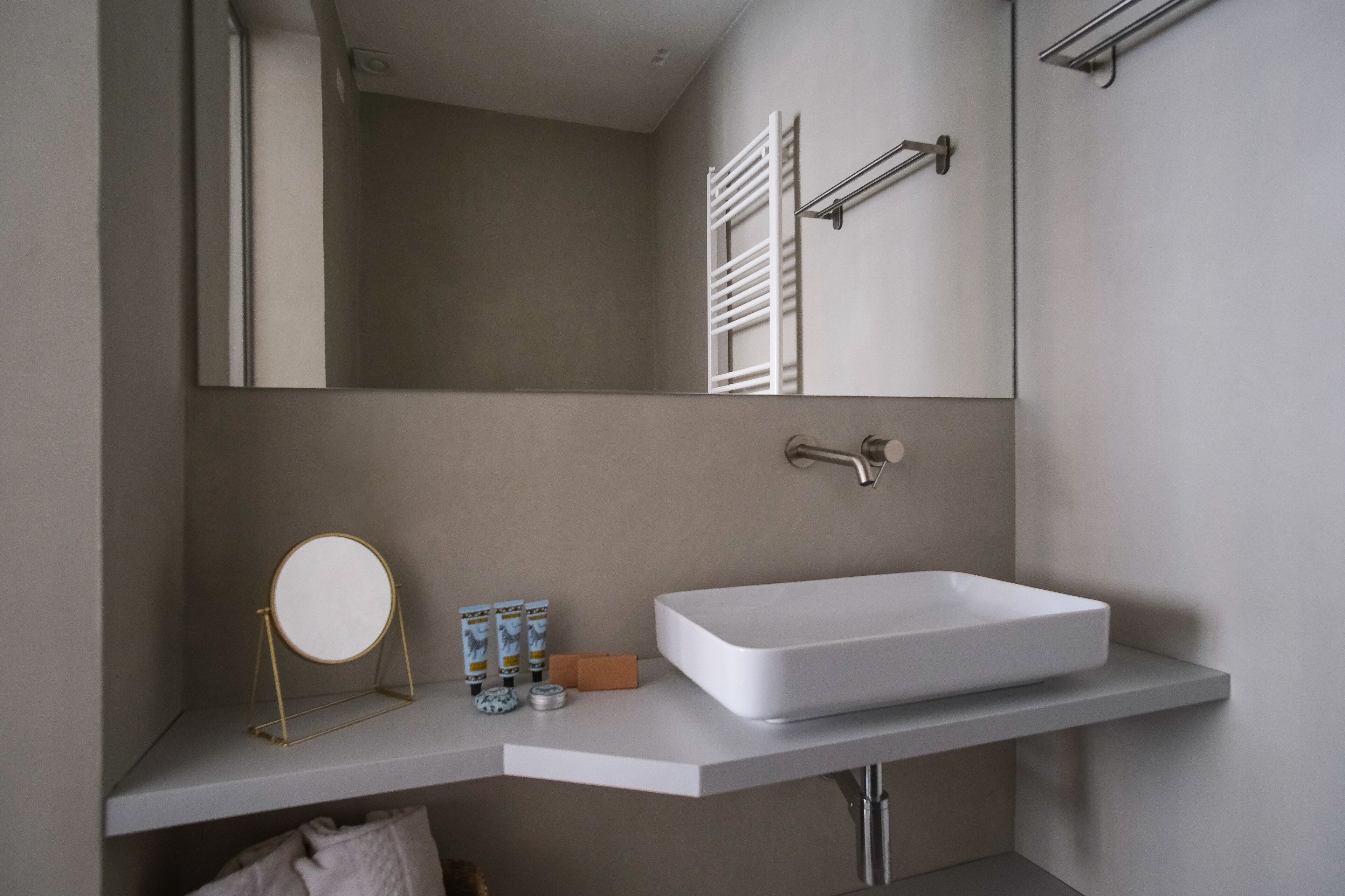 A modern bathroom with a large rectangular sink, a round mirror on a gold stand, and a towel rack above the sink.