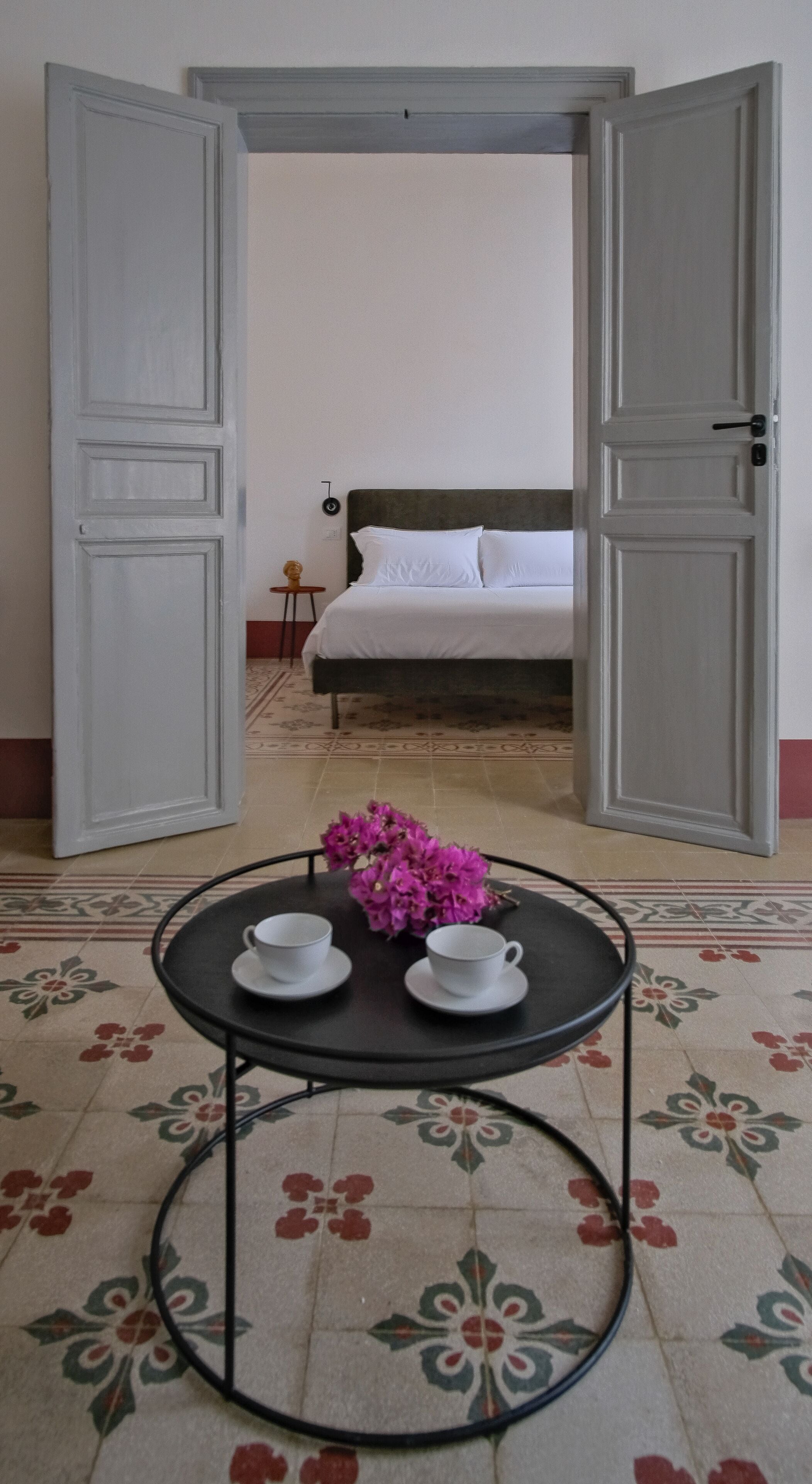 Un umbral abierto que da a un dormitorio con cama, una mesita con tazas y flores, y un suelo con diseño.