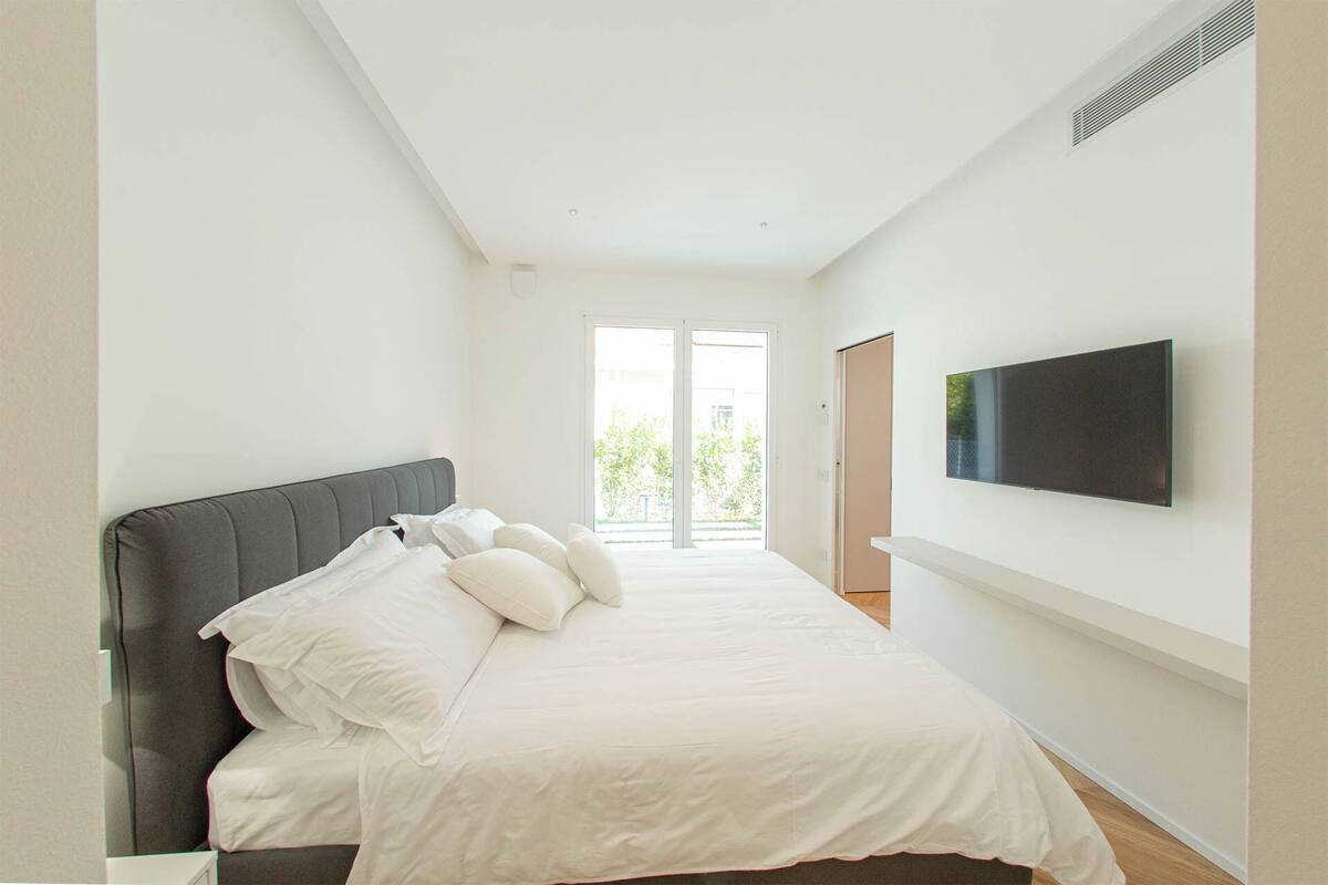 A bedroom with a bed, pillows, a TV mounted on the wall, and a sliding glass door leading to a balcony.