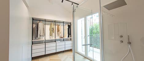A modern bathroom with a glass shower enclosure, a white vanity with drawers, and a clothing rack with various garments.