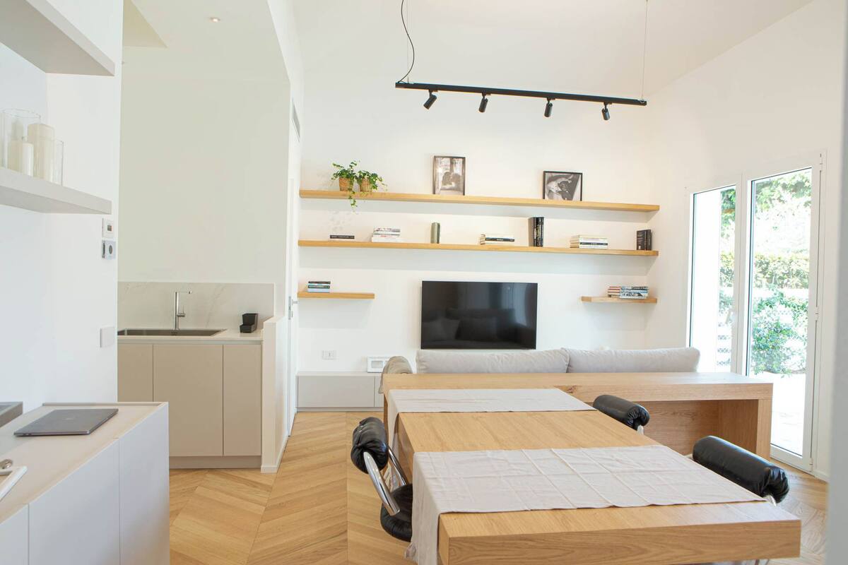 A modern kitchen with a built-in sink, a dining area with a wooden table and chairs, a sofa, and a TV mounted on the wall.