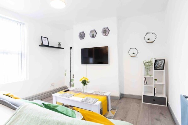 A modern living room with a flat-screen TV mounted on the wall, a white sofa, a small table with a vase of yellow flowers, and a white shelving unit with books and decorative items.