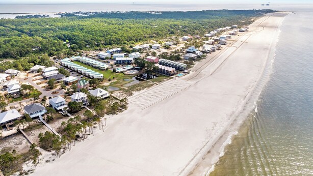 Vista aérea de un complejo turístico costero con propiedades frente al mar, una playa de arena y una zona boscosa.