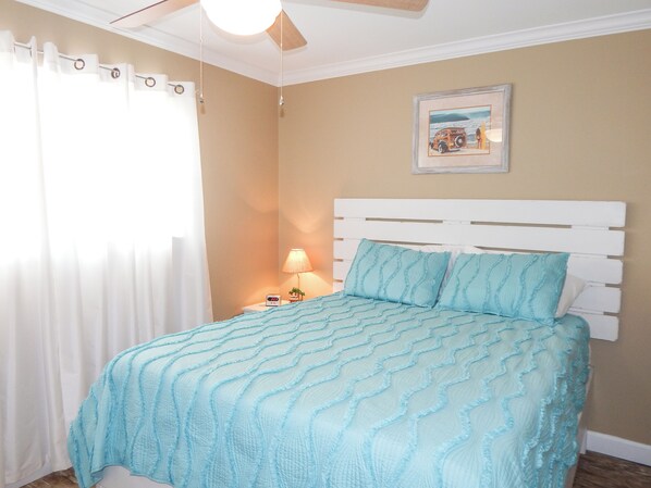 Un dormitorio con cabecera blanca, colcha azul con textura, mesita de noche con lámpara y un cuadro enmarcado en la pared.