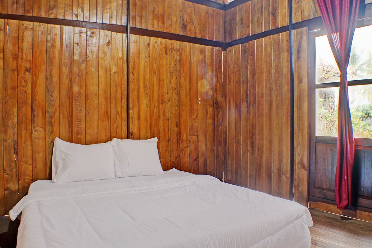 A room with wooden walls, a large bed with white bedding, and a window with a red curtain.