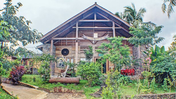 Un bâtiment traditionnel en bois, doté d’un toit en tuiles, entouré d’une végétation luxuriante et bordé d’un sentier en pierre.