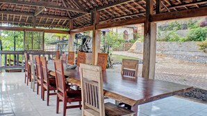 Un espace extérieur abrité, aménagé pour les repas, avec des tables et des chaises en bois, un sol carrelé et une vue sur un jardin et des bâtiments en arrière-plan.