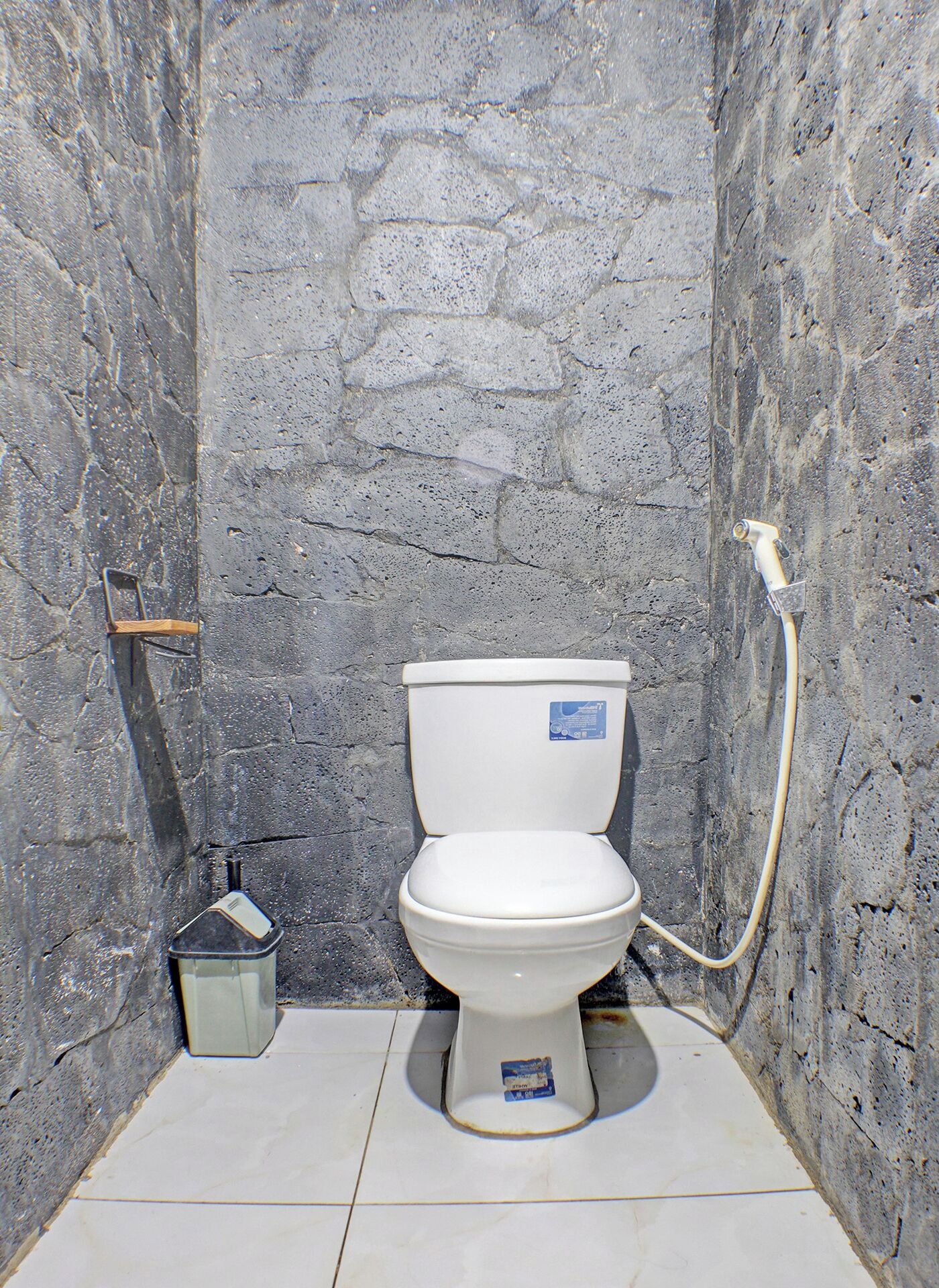 A small, stone-walled bathroom with a white toilet and a shower hose.