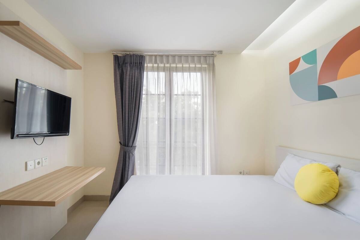 A hotel room with a bed, a TV mounted on the wall, a window with curtains, and a wooden shelf.