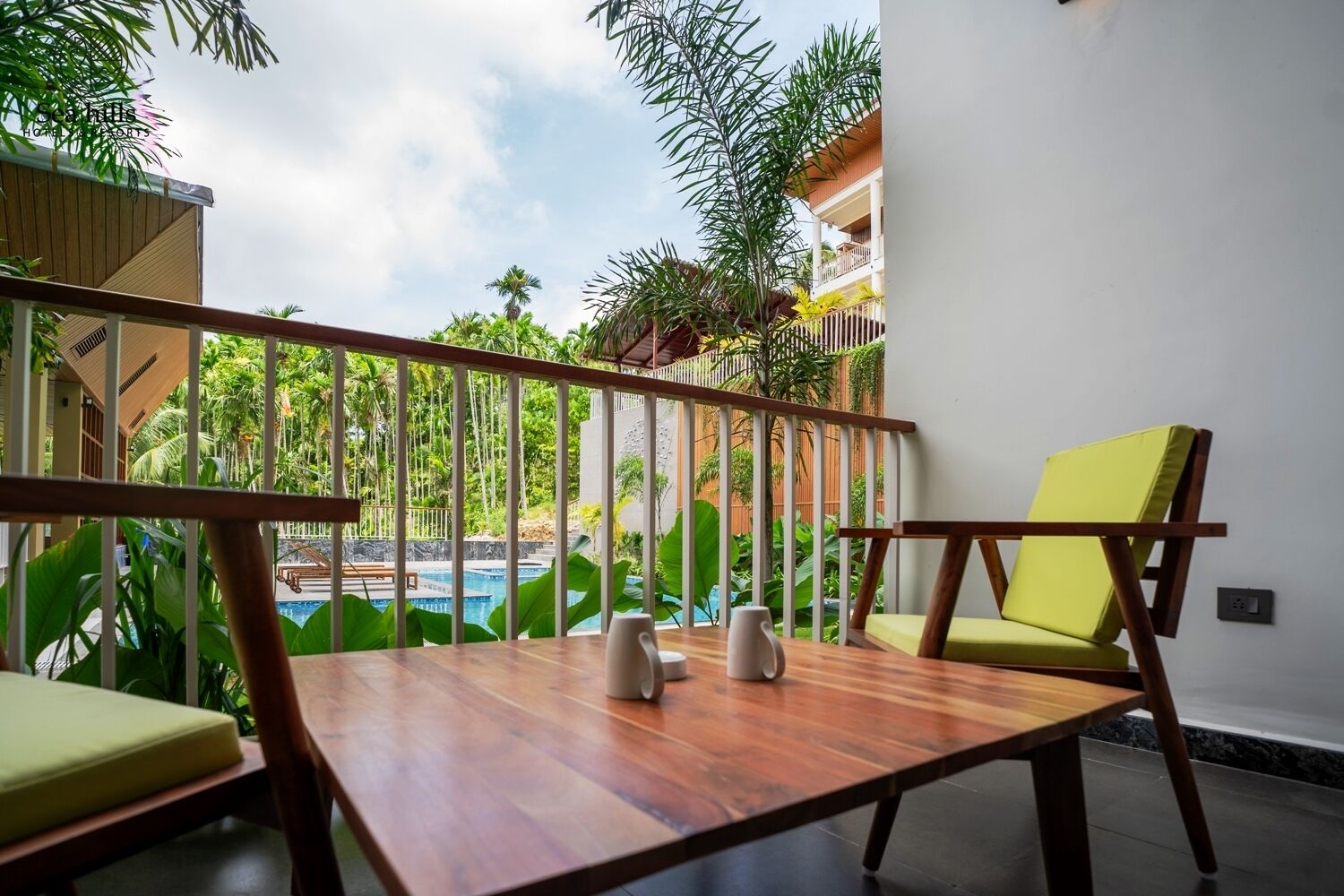 A balcony with wooden furniture, a pool view, and lush greenery.