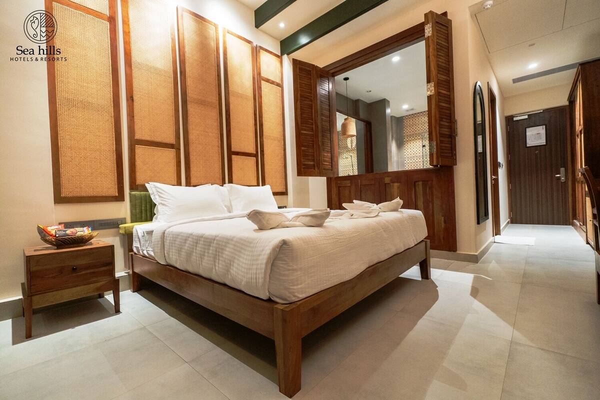 A hotel room with a large bed, wooden headboard, and a large mirror above the bed. There is a small wooden nightstand with a basket of snacks.