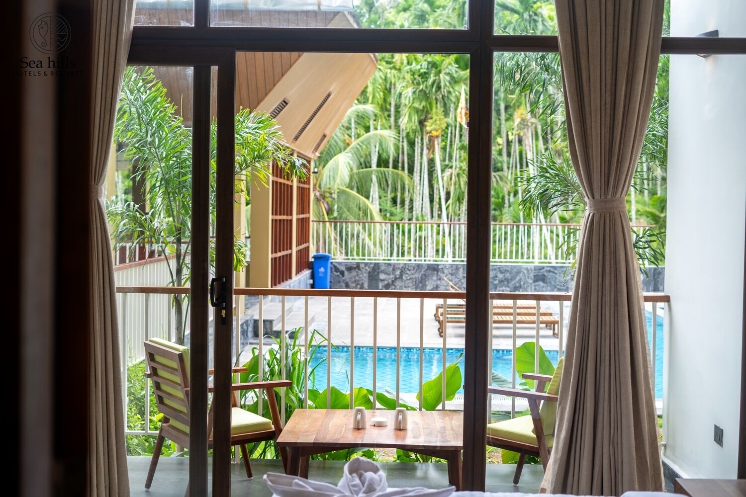 A room with a view of a pool, wooden furniture, and a tropical garden.