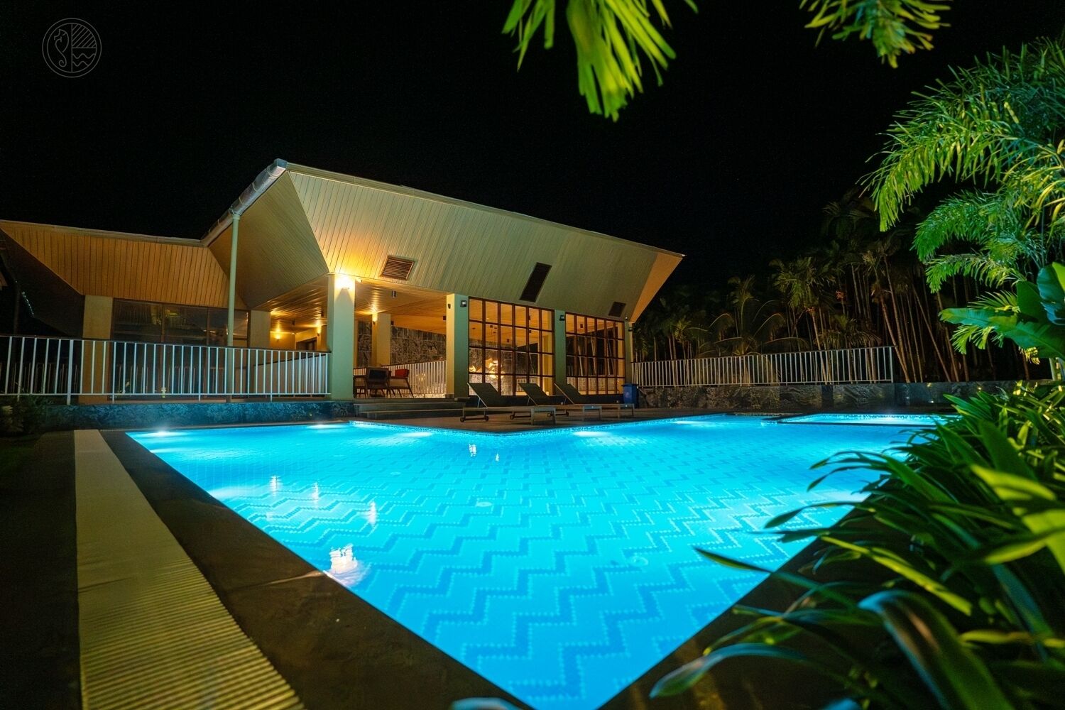 A swimming pool with a geometric tile pattern, surrounded by a white fence and lush greenery.