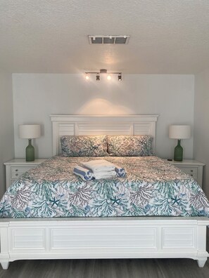 A bedroom with a white bed, patterned bedding, two bedside lamps, and a ceiling vent.