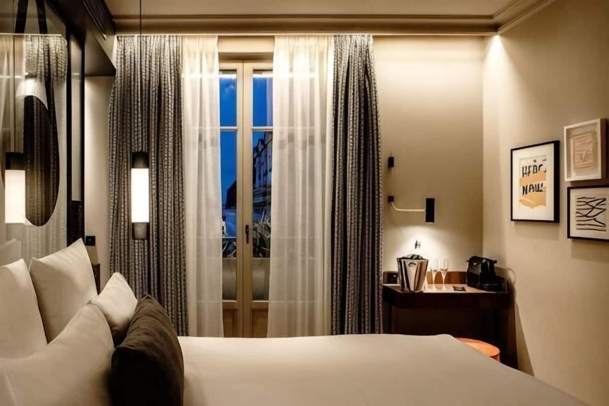 Habitación de hotel con cama, cortinas, escritorio con lámpara y cuadros en la pared.