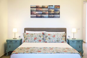 Een keurig opgemaakt bed met bloemenbedrukking, twee nachtkastjes met lampen, en een aan de wand hangend schilderij van een zonsondergang.