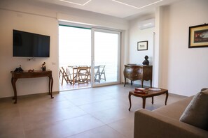 A living room with a flat-screen TV, a wooden coffee table, a sofa, a wooden side table, and a sliding glass door leading to a balcony with outdoor furniture.