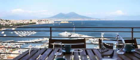 Una mesa con tazas vacías y una jarrón de vidrio, con vista a un puerto deportivo con barcos y una montaña distante.