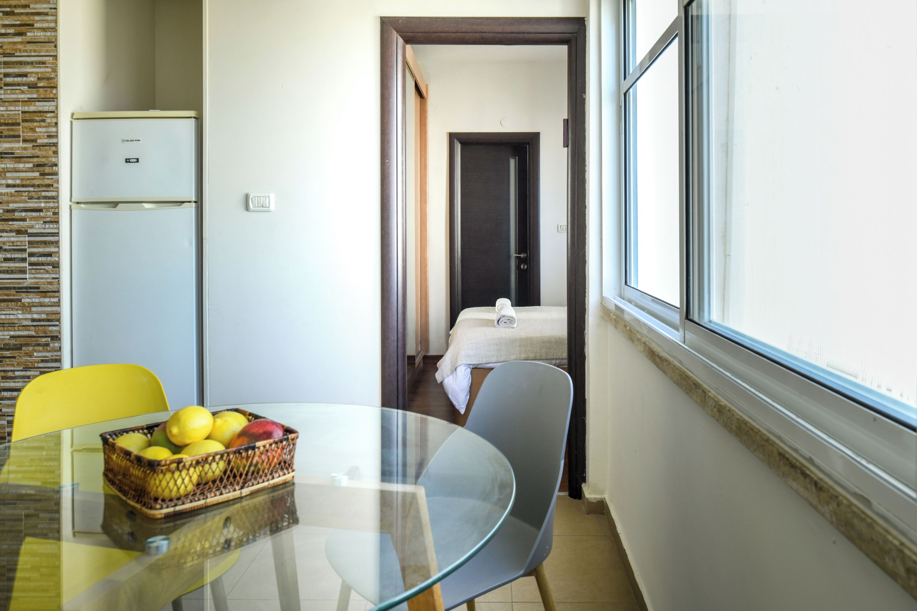 A hotel room with a bed, a small table with a fruit basket, a chair, and a refrigerator.