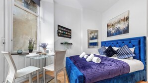 A modern bedroom with a blue upholstered bed, a glass desk, and a white chair. - Immaculate 1-bed Studio in London (London)