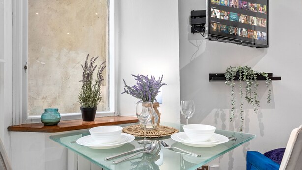 A dining area with a glass table set for two, a vase with purple flowers, a small potted plant, and a mounted TV displaying a movie. - Immaculate 1-bed Studio in London (London)