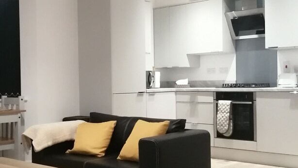 Una sala de estar moderna con un sofá negro, un banco y un tapete estampado. Se aprecia una cocina con armarios blancos y una estufa al fondo.